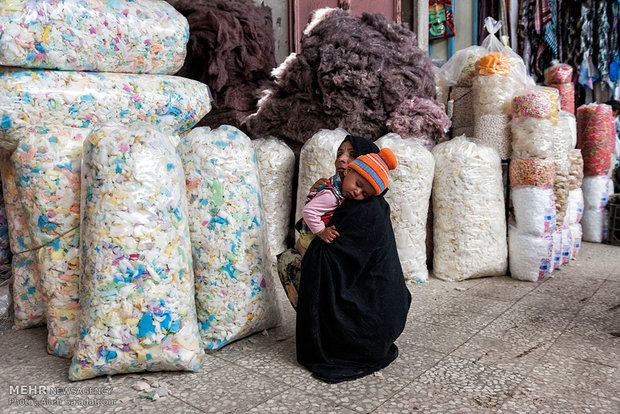 کودکی که از خستگی بر شانه مادرش خوابیده، مادر بیرون از مغازه تشک دوزی فروشی منتظر بستگان خود نشسته است.