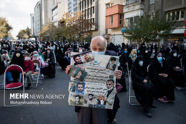 یک مرد مسن حاضر در مراسم یوم الله ۱۳ آبان در تهران تصویر مقام معظم رهبری ، شهید حاج قاسم سلیمانی و چند تن دیگر از شهدای ایران را در دست دارد