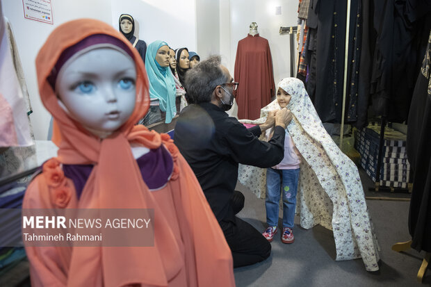 یک پدر در حال پوشاندن چادر  به فرزند خود در یکی از غرفه های عرضه چادر در نمایشگاه عفاف و حجاب است