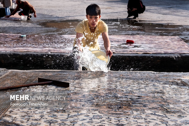 پسری در حال آب ریختن از چشمه علی بر روی فرش برای شست شوی آن است