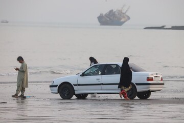 An Iranian family on the northern shore of the Strait of Hormuz, with a disabled ship visible in the background.